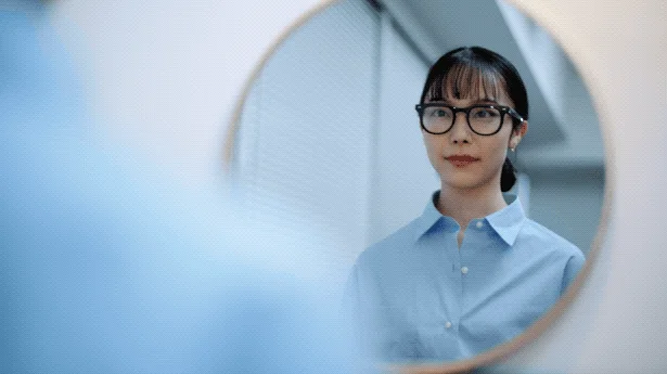 青いシャツと黒縁眼鏡をかけた若いアジア人女性が、丸い鏡に映る自分の姿を見つめている