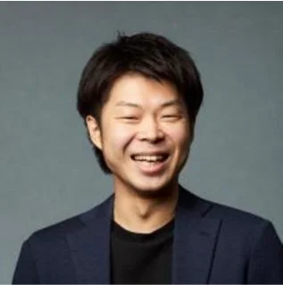 笑顔でカメラを見つめる東洋系の男性のポートレート。紺色のジャケットに黒いシャツを着用し、親しみやすい印象を与えています。