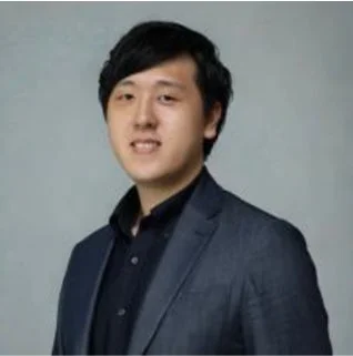 グレーの背景を背にした、スーツを着た若いアジア人男性のポートレートです。男性はカメラに向かってわずかに微笑んでおり、プロフェッショナルな印象を与えます。