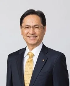 濃い色のスーツと黄色いネクタイを着用し、眼鏡をかけた男性が笑顔で写っているポートレート