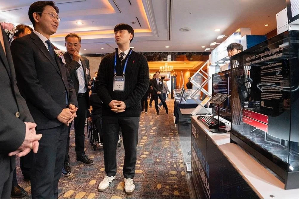 テクノロジーイベントで、男性がAIコンピューティングシステムを説明している様子です。スーツ姿の参加者たちが展示ブースでデモンストレーションを見学しています。