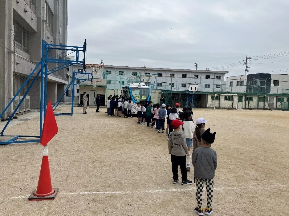 校庭で子供たちが列を作り、バスのような乗り物に乗ろうとしている様子が写っています。