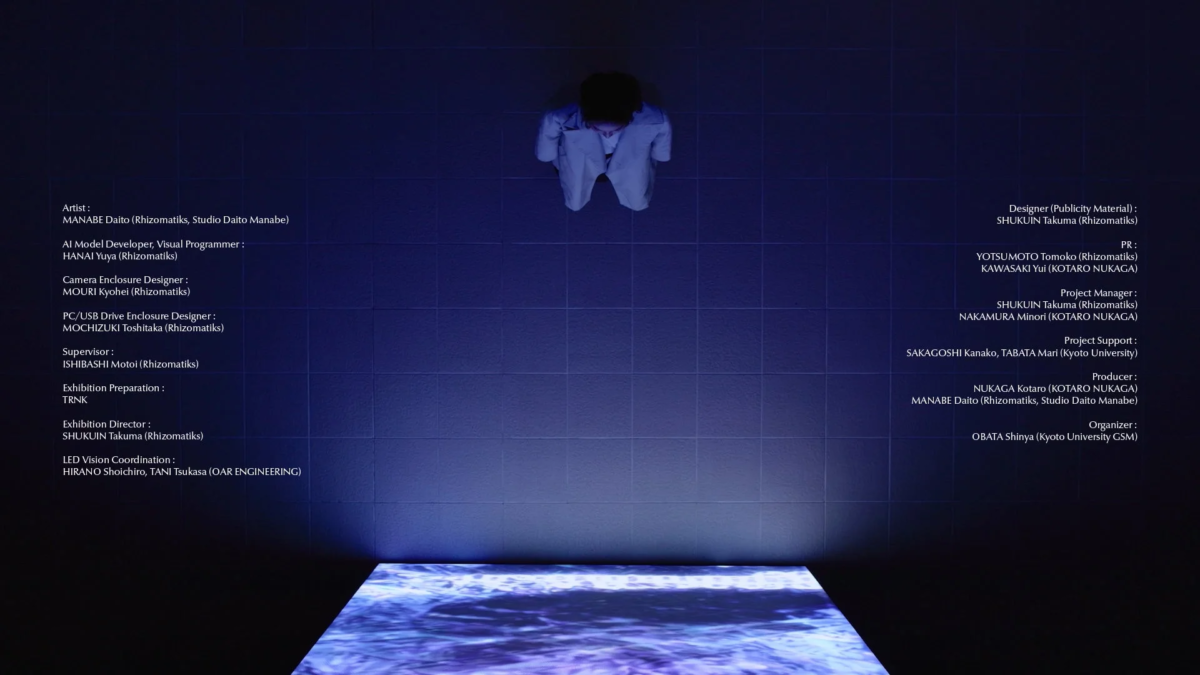 暗い空間で一人の人物が青い光の下に立ち、足元にある水面が波打つようなデジタルディスプレイを見下ろしている様子