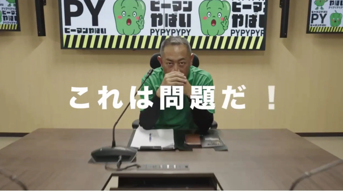 会議室で深刻な表情の男性と、ピーマンのキャラクターが描かれたテレビ画面