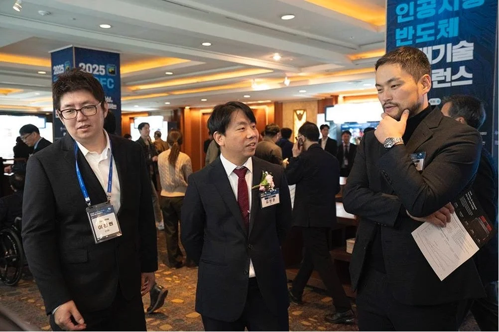 会議会場のような場所で、スーツを着た3人の男性が会話したり、周囲を見渡したりしている様子が写っています。背景にはAIや半導体技術に関すると思われるバナーが見えます。