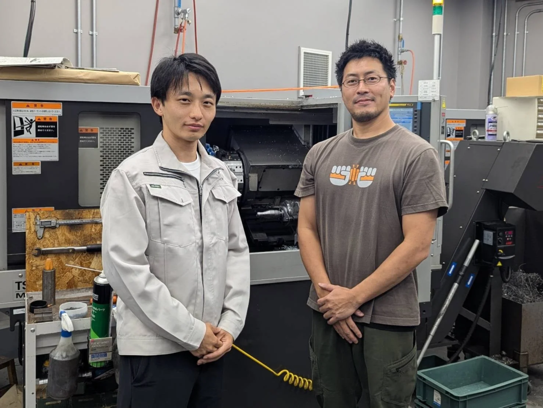 工場のような場所で、2人の男性が機械の前で立っています