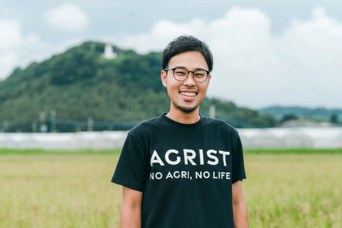 緑豊かな畑に立つ、笑顔の若い男性のポートレートです。彼は「AGRIST NO AGRI, NO LIFE」と書かれた黒いTシャツを着ており、背景には山と曇り空が広がっています。