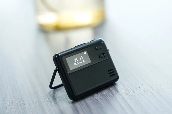 時刻と日付が表示された小型の黒いデジタルデバイスが、木目調のテーブルの上に置かれている様子
