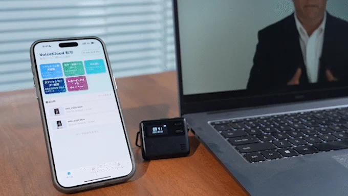 スマートフォンに音声文字起こしアプリ「VoiceCloud 転写」が表示され、その隣に小型レコーダーとラップトップが置かれています。