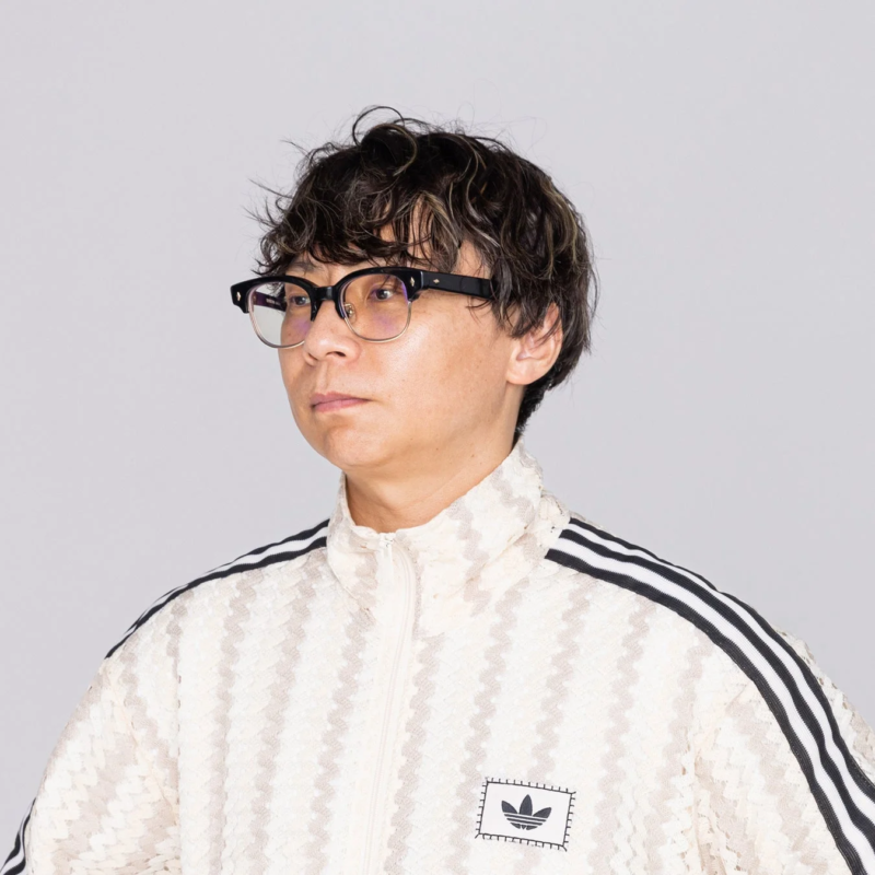 眼鏡をかけた男性が、アディダスのクリーム色のトラックジャケットを着用し、白い背景の前で横向きに立っているポートレート写真