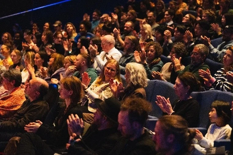 薄暗い会場で、多くの観客が拍手をしている様子が写っています。