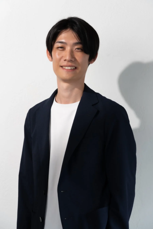 白背景の前で、紺色のブレザーと白Tシャツを着用した若い日本人男性が笑顔でポーズをとっているポートレート写真です。