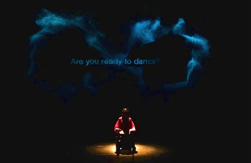 暗い舞台の中央で車椅子に座る人物と、その上部に青い光で「Are you ready to dance?」というメッセージが映し出されている様子。