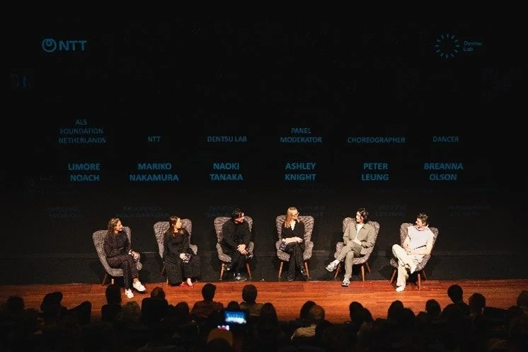 舞台上で6名の登壇者が座り、背景のスクリーンには名前と役職が映し出されています。