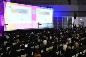 広い会場で大勢の聴衆が座ってプレゼンテーションを聞いている様子