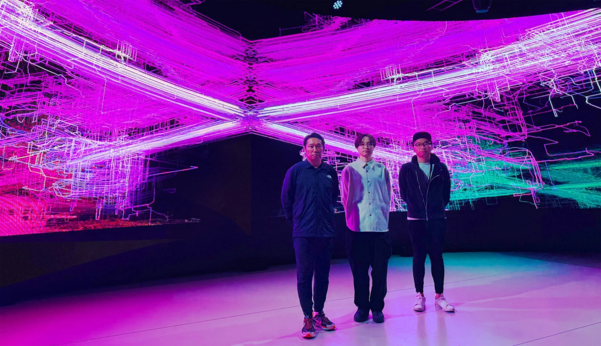 未来的なデジタルアートが映し出されたステージに3人の男性が立っています