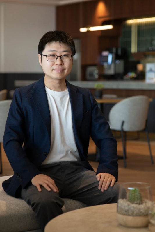 眼鏡をかけたアジア人男性が、ネイビーのジャケットと白いTシャツを着てソファに座り、カメラに向かって微笑んでいるポートレートです。