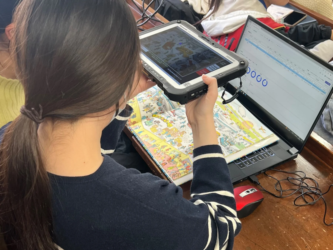 一人の人物が、頑丈なタブレット(Panasonic Toughbook)を『ウォーリーを探せ』のような絵本にかざしている様子を後ろから捉えた画像です。タブレットの画面には絵本の内容が映し出されており、隣にはデータが表示されたノートパソコンも開かれており、デジタルとアナログを組み合わせた学習や研究の場面を示唆しています。