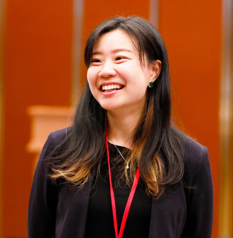 笑顔のアジア系女性のポートレートです。彼女は黒いジャケットを着用し、首には赤いランヤードを下げています。屋内でのイベントや会議のような状況を思わせる、明るく親しみやすい雰囲気の画像です。