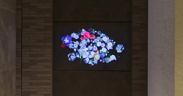 壁に設置されたディスプレイに、様々な色合いの花々が抽象的な形状で描かれたデジタルアートが表示されています。青、白、ピンク、赤などの花が暗い背景に浮かび上がり、空間を華やかに彩っています。