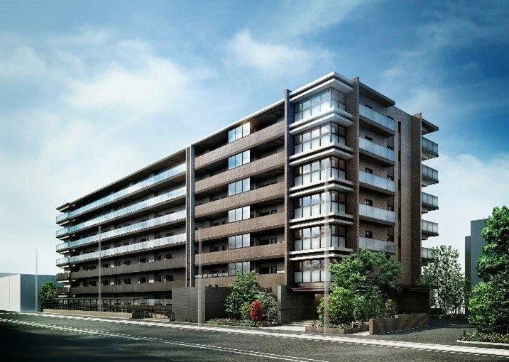 青い空の下に建つ、現代的なデザインのマンションの外観写真です。複数のバルコニーと大きな窓が特徴で、周囲には緑豊かな植栽と道路が見えます。新築または比較的新しい集合住宅のようです。