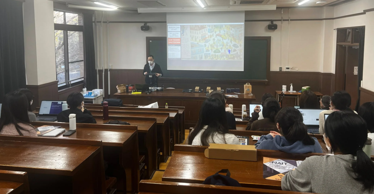 大学の講義室で、講師がプロジェクターを使ってプレゼンテーションを行っています。複数の学生がノートパソコンを開いて授業を受けている様子です。