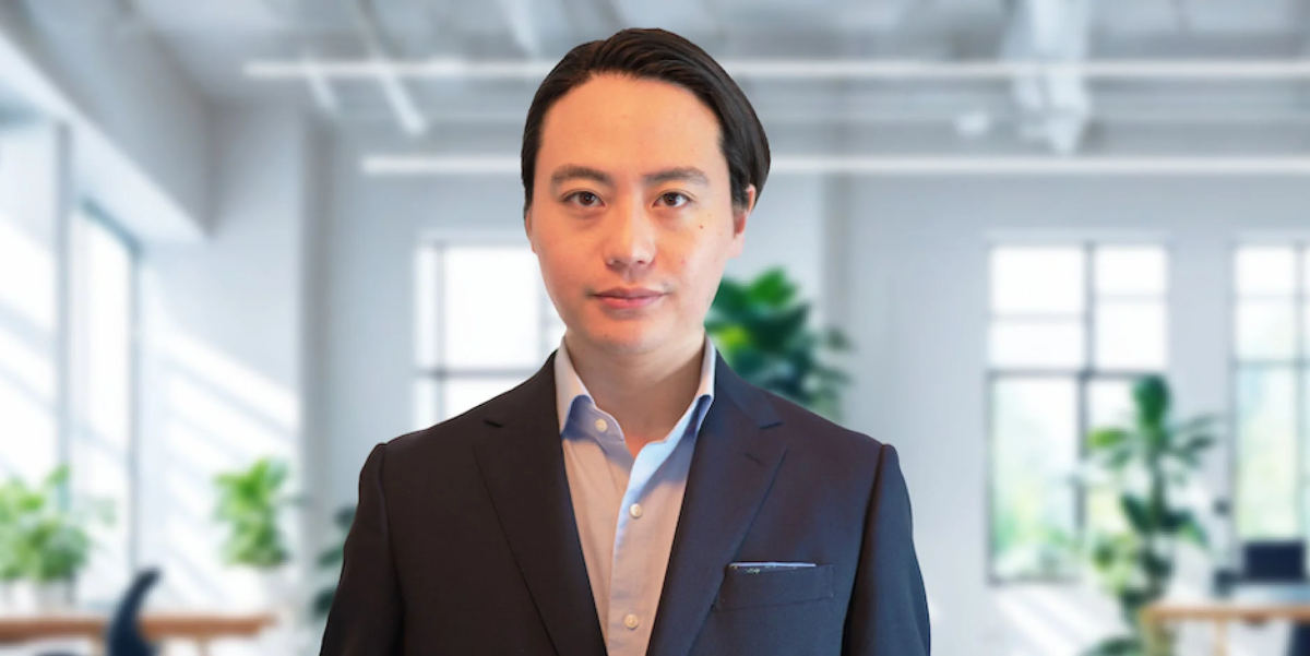 ビジネススーツを着用したアジア系の男性が、ぼやけたオフィスを背景にカメラを見つめているポートレート写真です。