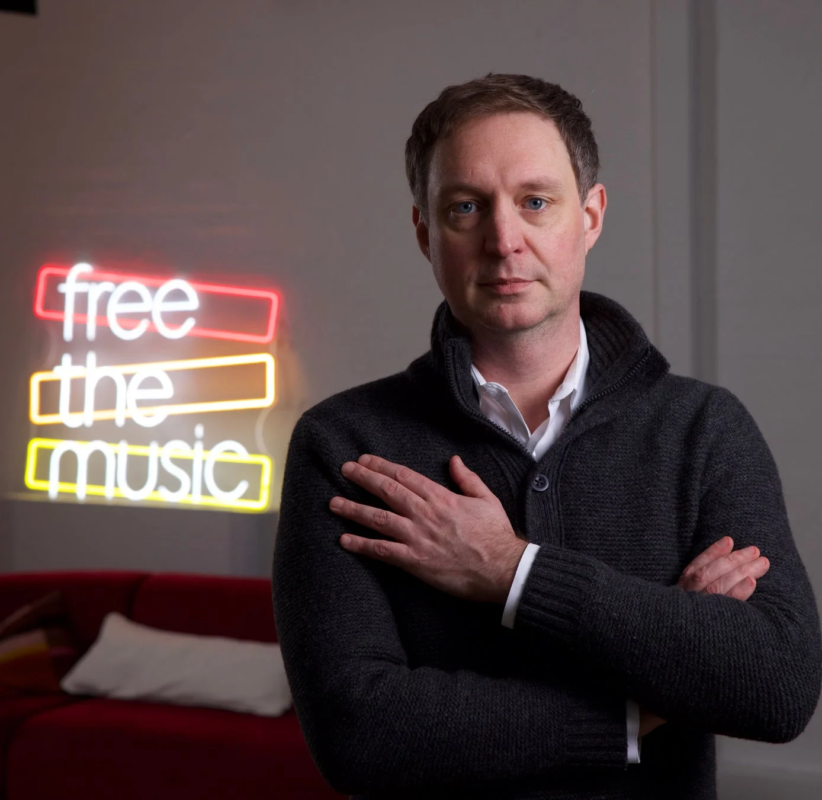 「free the music」と書かれたネオンサインを背景に、ダークグレーのカーディガンを着た男性が腕を組み、真剣な表情でカメラを見つめているポートレートです