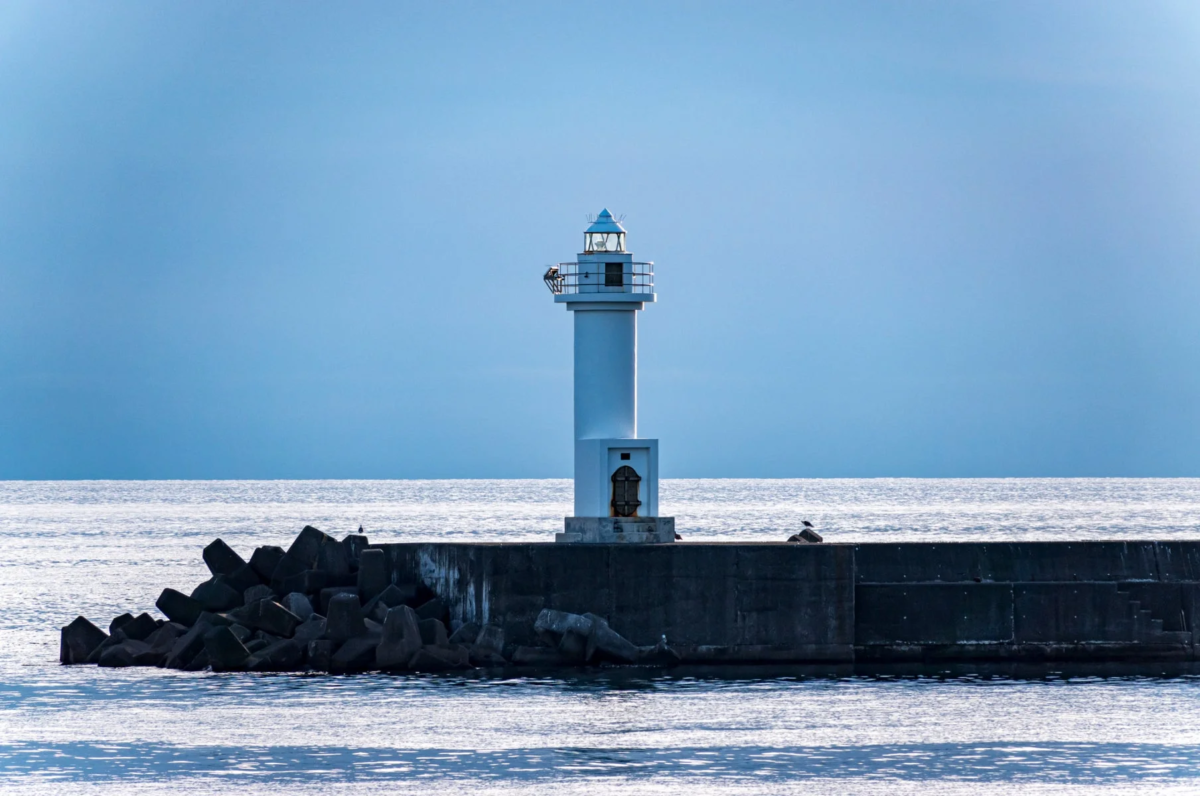 青い空と海を背景に、白い灯台が防波堤の上に立つ風景。水面には光が反射し、テトラポッドが波打ち際を囲んでいます。