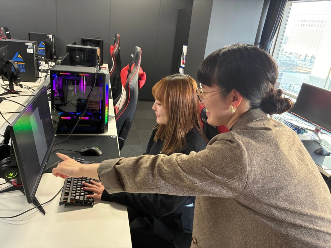 eスポーツの専門学校のような教室で、一人の女性が別の女性にゲーミングPCの画面を指差しながら説明している様子