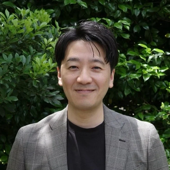 緑豊かな背景を背に、笑顔でカメラを見つめるアジア系の男性のポートレート写真です。グレーのジャケットを着用しています。