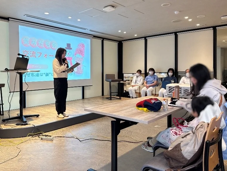 交流フォーラムで発表する女性。テーマは「なぜ私たちは“顔”に」