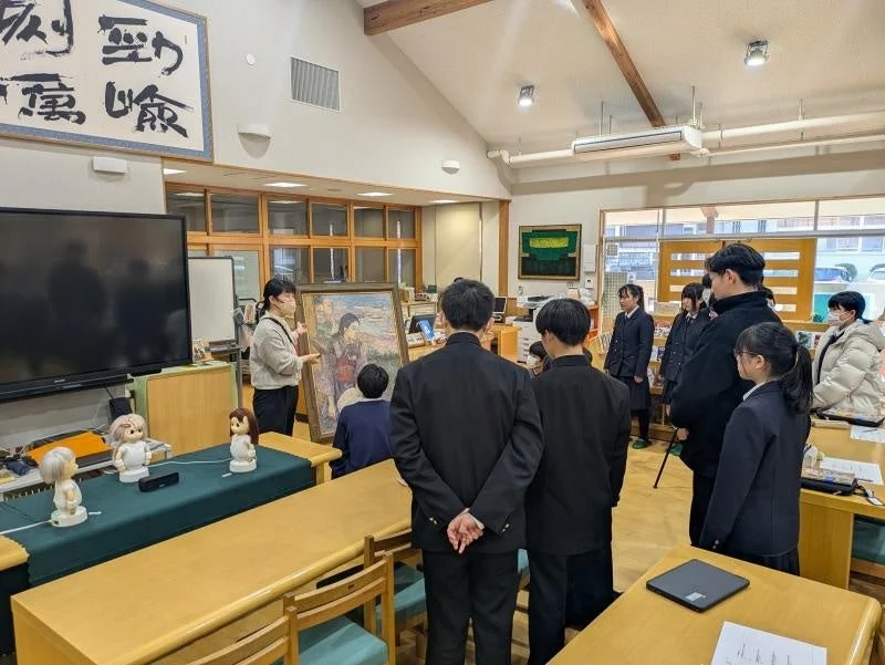 学校の教室で、先生が制服を着た生徒たちに絵画を提示し説明している。生徒たちは絵に注目し、学びの場となっている。机の上には小型ロボットも複数置かれ、多様な学習が行われている様子