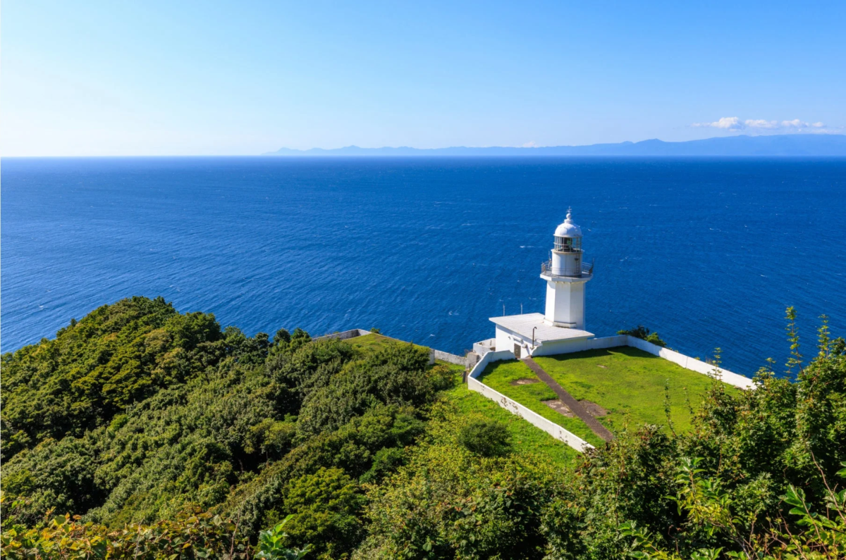 青い海と青い空が広がる岬に立つ白い灯台の風景。緑豊かな丘と白い灯台のコントラストが美しい、晴れた日の絶景です。