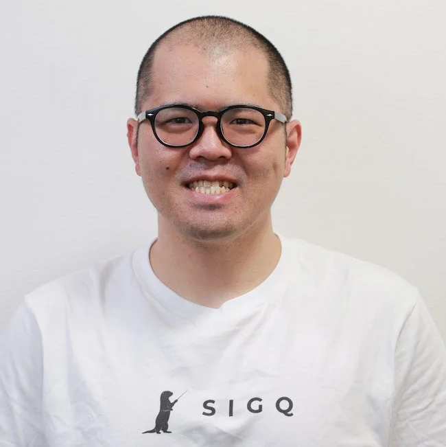 白いTシャツを着て眼鏡をかけた男性が、笑顔でカメラを見つめているポートレート写真です。Tシャツには動物のイラストと「SIGQ」というロゴがプリントされています。