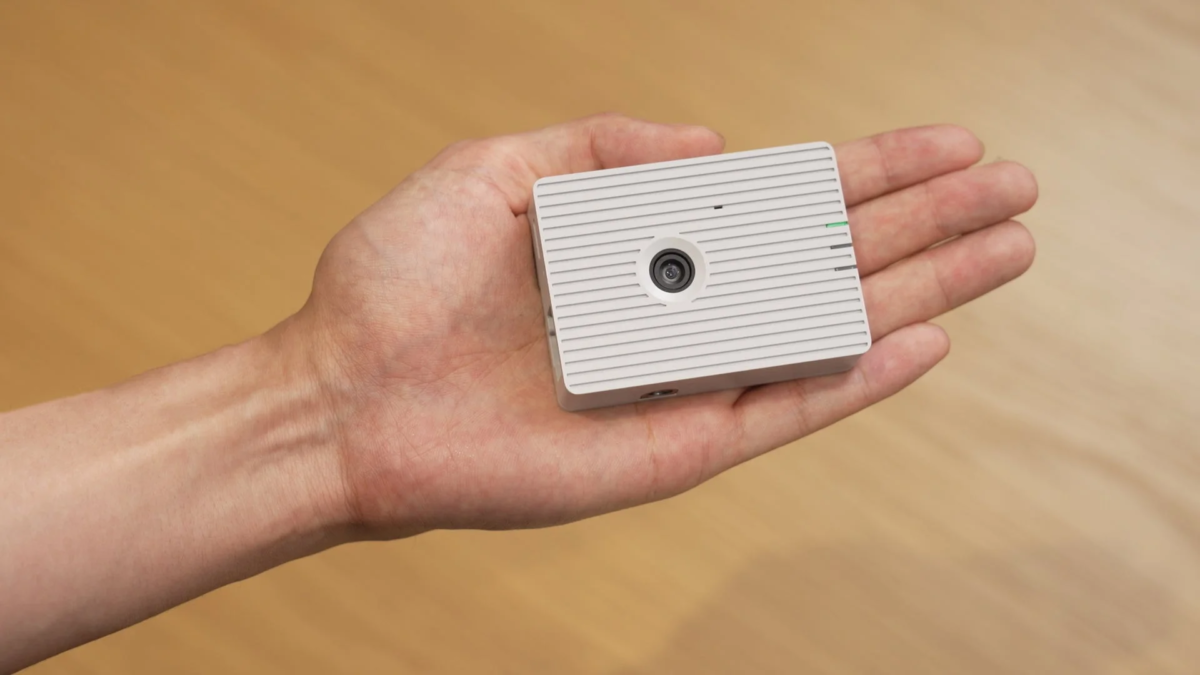 手のひらに乗せられた小型の白い電子デバイスのクローズアップ。中央にカメラレンズがあり、表面には横縞模様のデザインが施されています。