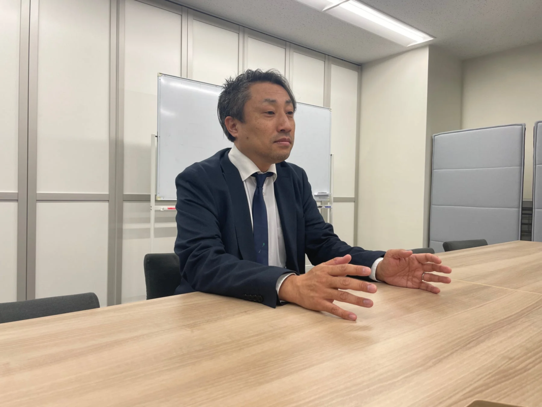 会議室の木製テーブルに座り、スーツ姿の男性が真剣な表情で話しており、両手を広げてジェスチャーを交えています。ビジネスシーンでの打ち合わせや説明の様子を捉えた画像です。