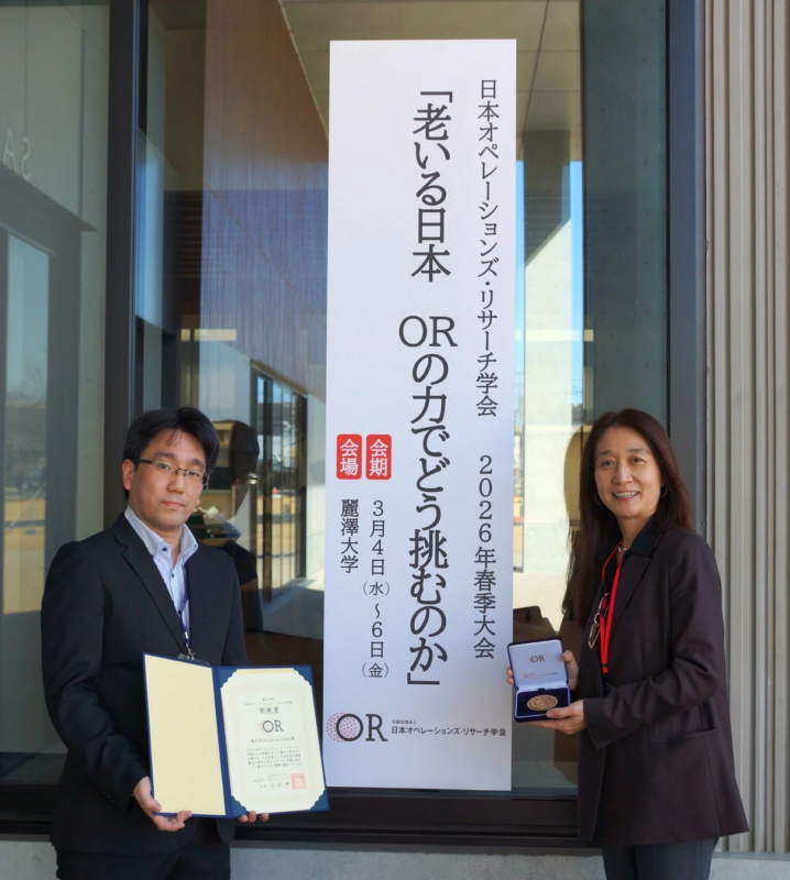 日本オペレーションズ・リサーチ学会の2026年春季大会のバナーの前で、男性が賞状を、女性がメダルを持って立っています。学会での功績を称える表彰式の一場面と見られます。
