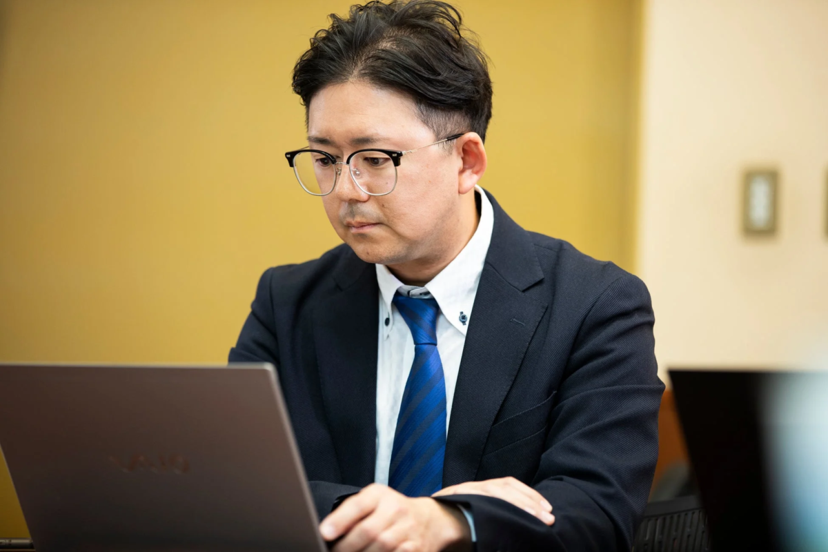 スーツとネクタイを着用しメガネをかけた男性が、ノートパソコンを真剣な表情で見つめて作業している様子。オフィスでのビジネスシーンを捉えた一枚です。